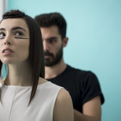 ragazza con capelli nera e canotta bianca viene pettinata da parrucchiere con t-shirt nera