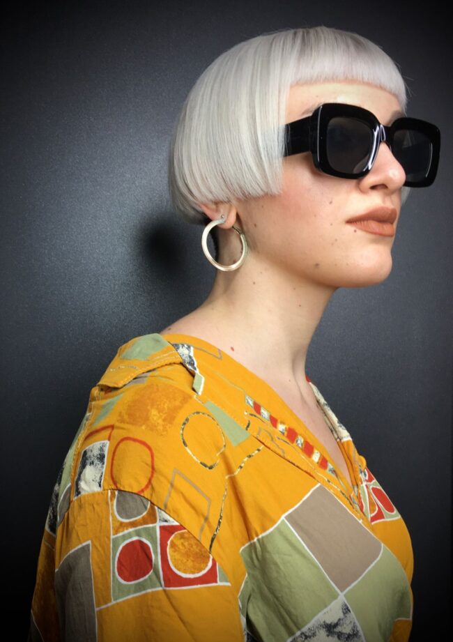 ragazza con caschetto corto e frangia bianchi decolorato, occhiali da sole neri e camicia gialla
